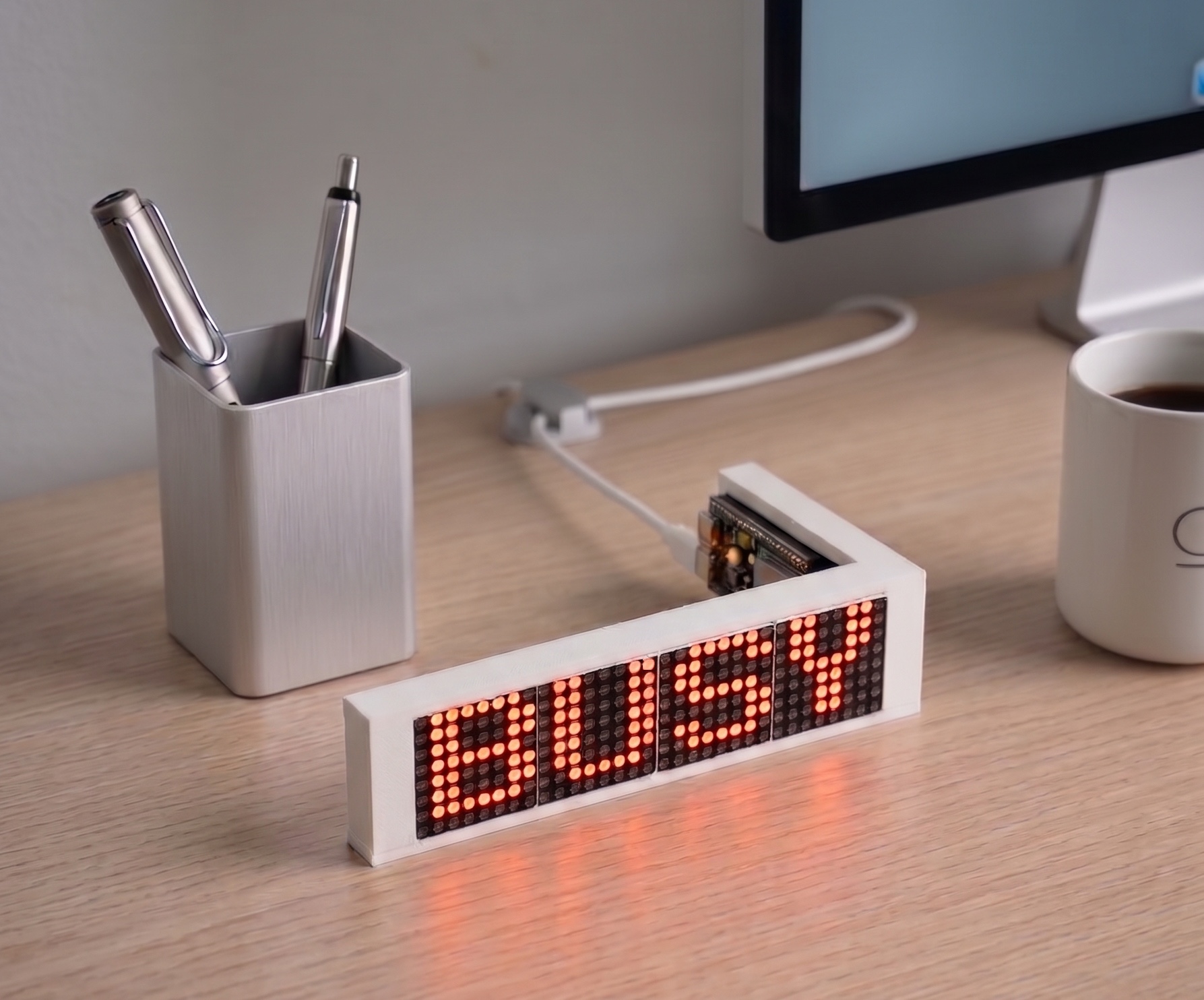 The LED Ticker on a desk in its white frame, its red dot-matrix display reading BUSY, connected to an ESP32-S3 board over a white USB-C cable.